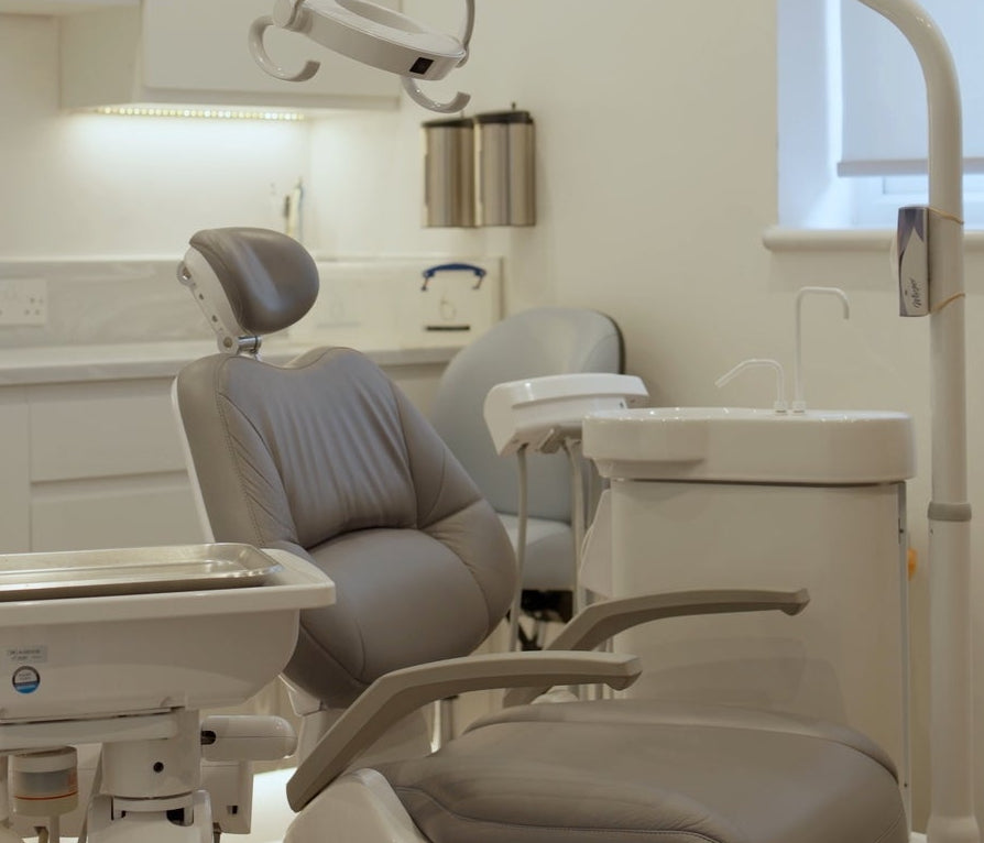 A modern dental office interior with a gray dental chair, white dental arm, sink, cabinets, and a window, viewed through an open doorway.