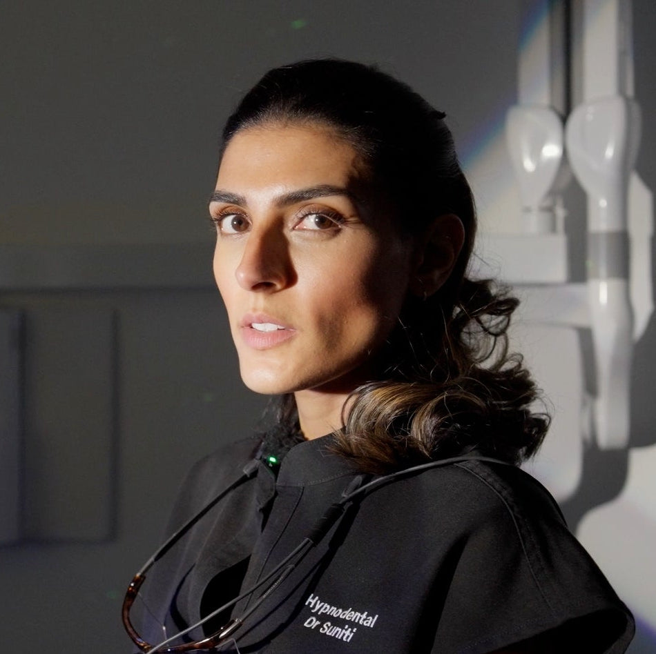 Dr Suniti in contrasting light in black medical scrubs stands in profile against a dim gray wall, with a stethoscope around her neck and white medical equipment visible behind her.