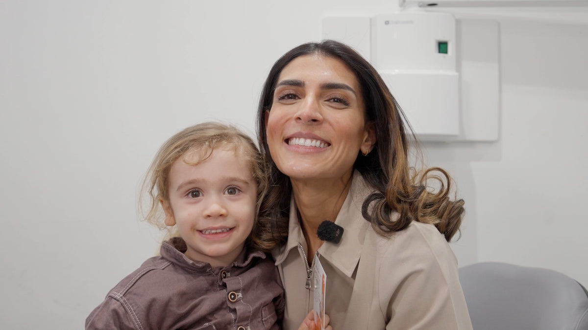 Dr Suniti and a young dental patient with light hair, both wearing brown and beige clothing, standing indoors against a light gray wall.