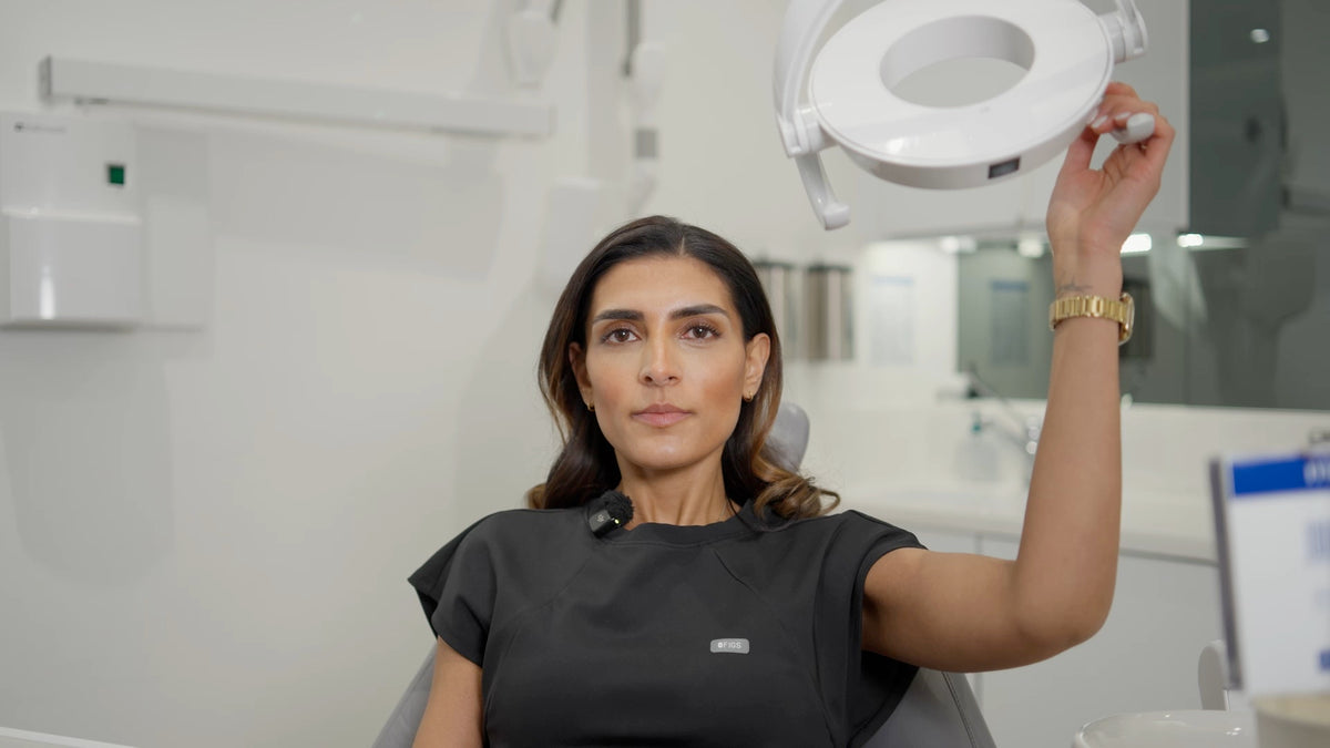 Dr Suniti in a black scrub top sits in a dental chair holding up a white dental operating light in a bright, modern dental office.