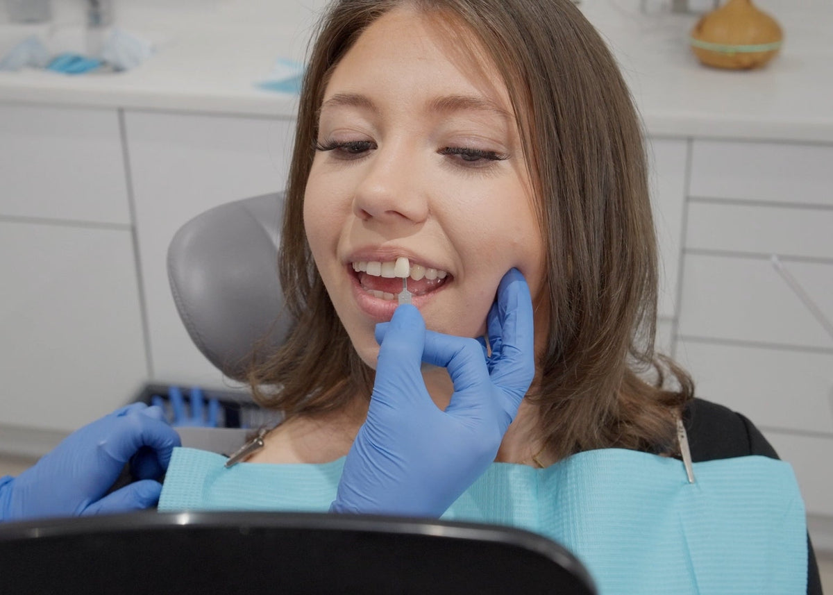 A woman sitting in a dental chair with a light blue bib, having her teeth examined by a dentist wearing blue gloves in a bright dental office.